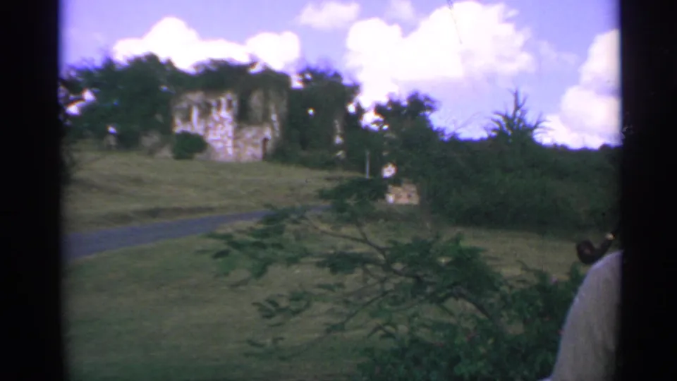 1967 home-movie frame of castle-like ruins on a grassy hillside with tropical foliage and a partial traveler at right.