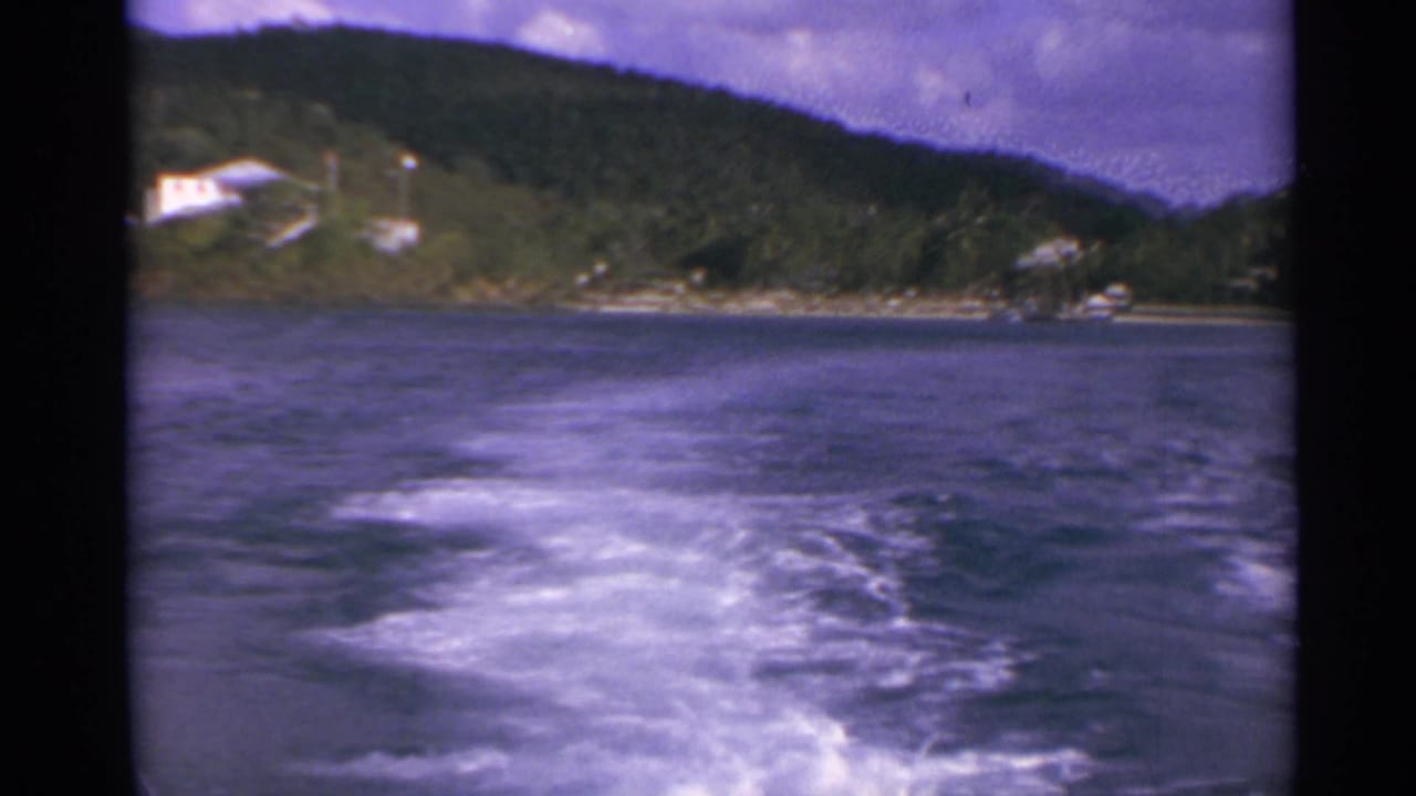1967 8mm view from a moving boat in the US Virgin Islands, with white wake, dark water, green hills, and small shoreline houses.