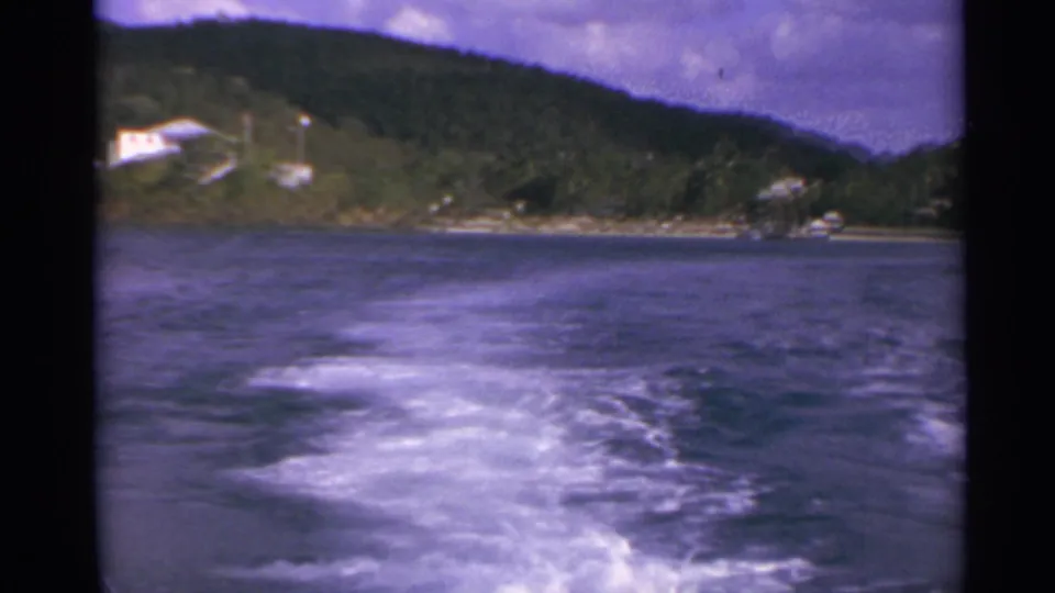 1967 8mm view from a moving boat in the US Virgin Islands, with white wake, dark water, green hills, and small shoreline houses.