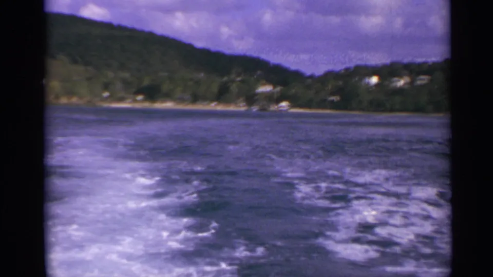Moving-boat view in the US Virgin Islands, showing foamy wake across open water beneath a cloudy sky and distant hillside homes.