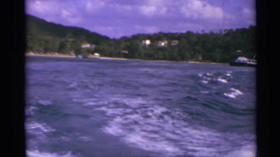 1967 home-movie frame from a boat, with rolling wake, wooded US Virgin Islands shoreline, scattered houses, and a small boat near shore.