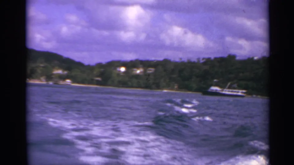 US Virgin Islands boat-ride frame from 1967, with choppy wake, purple-tinted sky, hillside vegetation, shoreline houses, and a small boat.