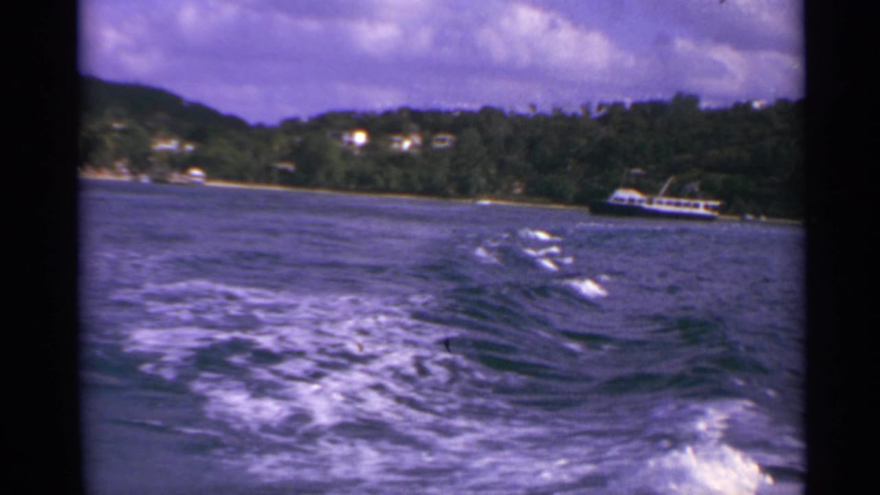 1967 8mm view from a moving boat in the US Virgin Islands, showing wake, cloudy sky, green shoreline, and a white vessel offshore.
