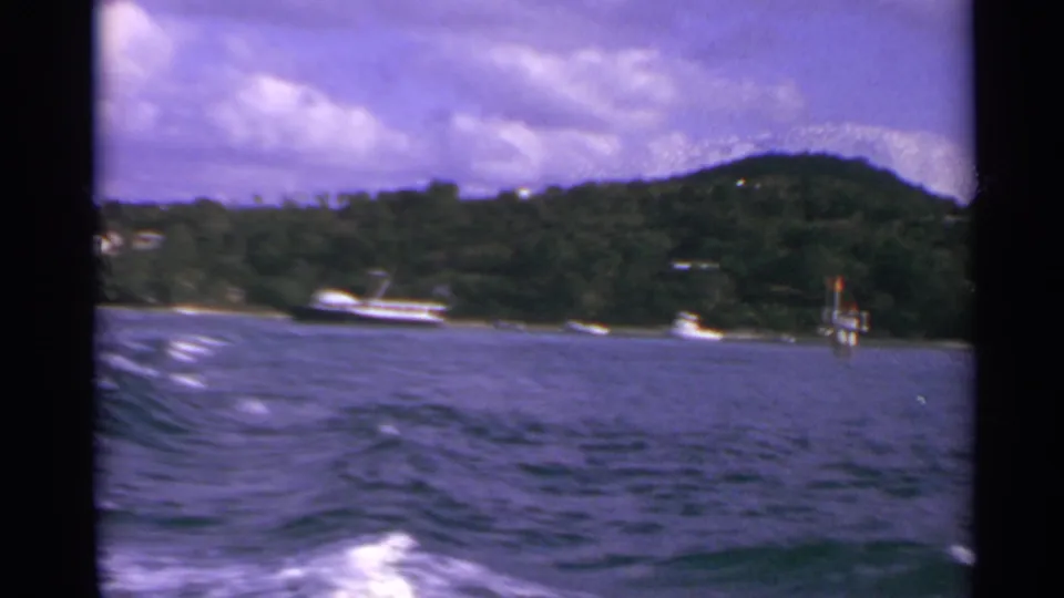 1967 US Virgin Islands home-movie frame showing calm water, small boats, a white vessel, and a wooded hillside under clouds.