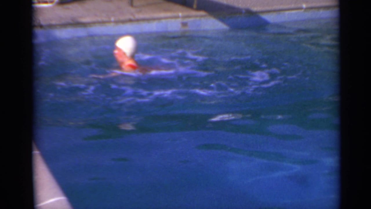 1967 8mm frame of a swimmer in a white cap and red swimsuit moving across a sunlit pool toward the deck.