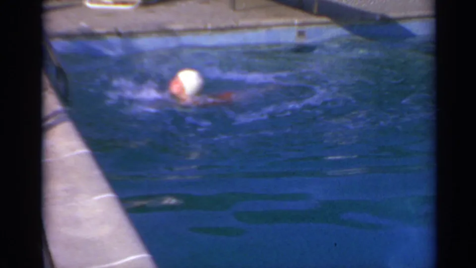 The swimmer remains partly submerged in blue water, with the pool deck and sunlit edge behind her.