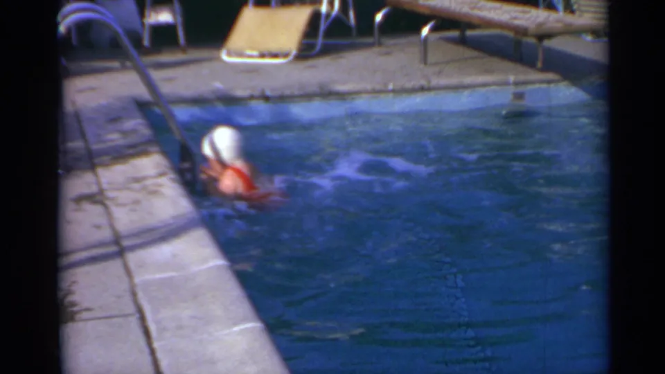 The swimmer is close to the pool ladder, with lounge chairs and the concrete pool deck visible in the background.