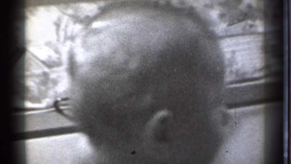 The baby's head fills the frame from behind, with bedding, a window area, and bed-frame lines visible in the 1939 home movie.
