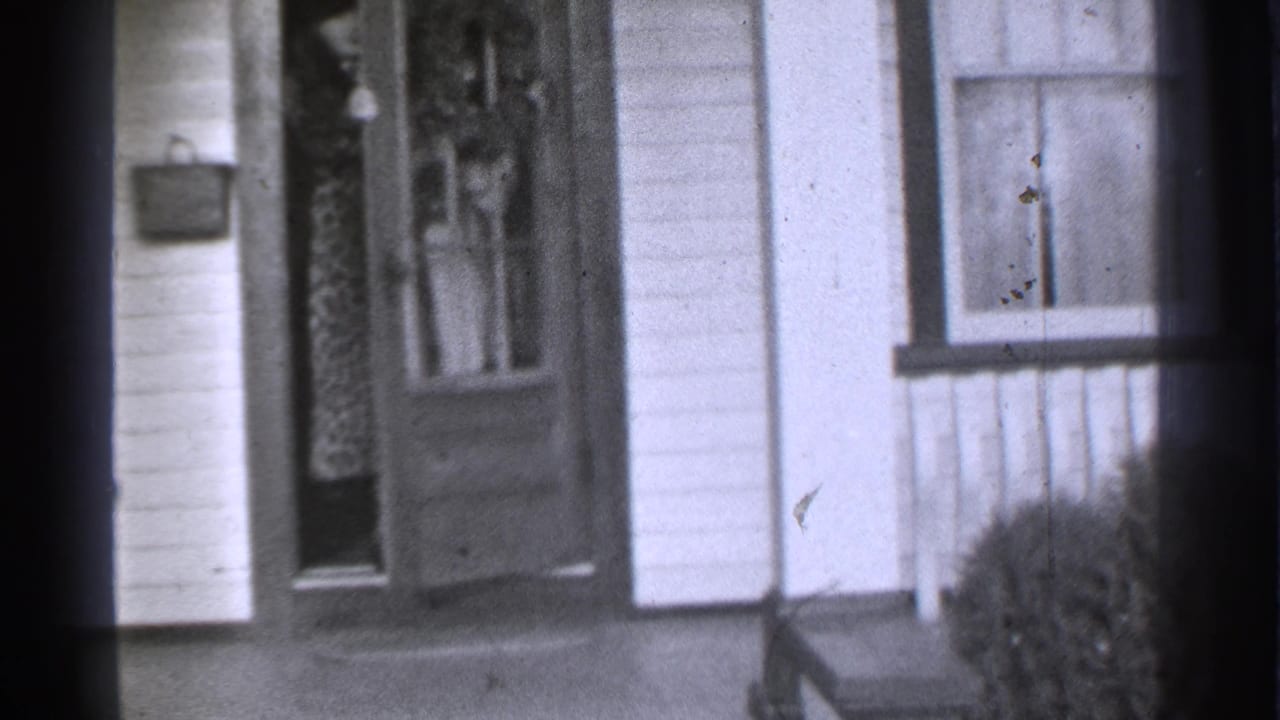 House entrance with front door, window, white siding, porch, and shrub at right.