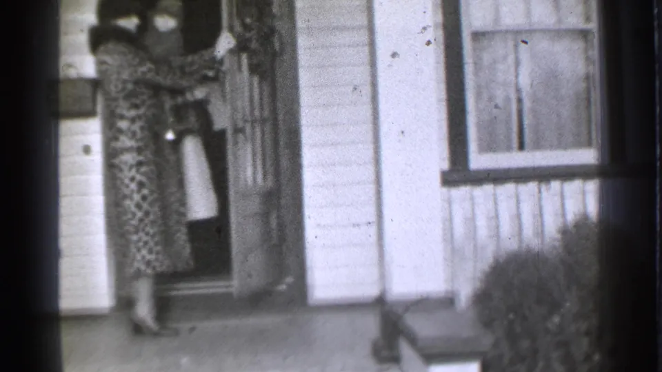 1939 Danielson, Connecticut frame showing a woman in a patterned dress and heels standing near the front door of a house.