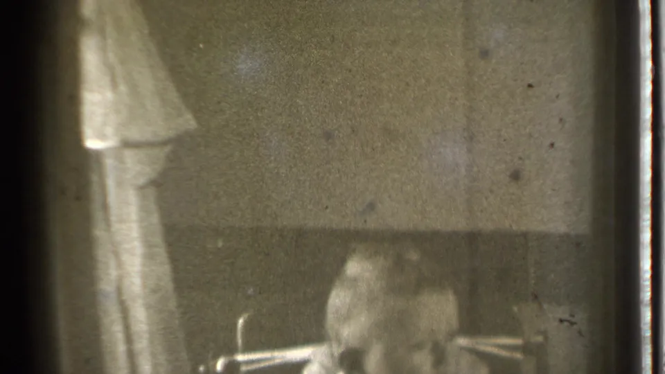 Shadowed home-movie interior from Danielson, Connecticut, with a light curtain and a partially visible seated person in soft focus.
