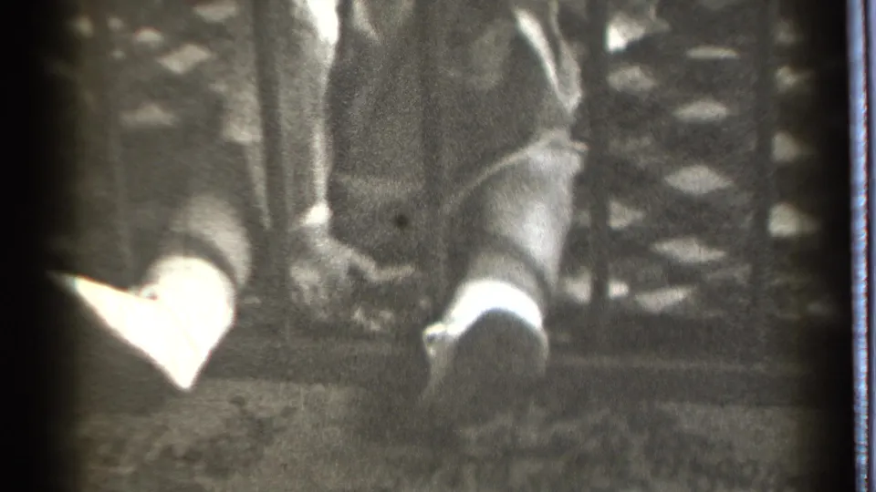 A grainy 1939 home-movie frame shows the child's lower body behind bars, with one light foot extended near fabric.