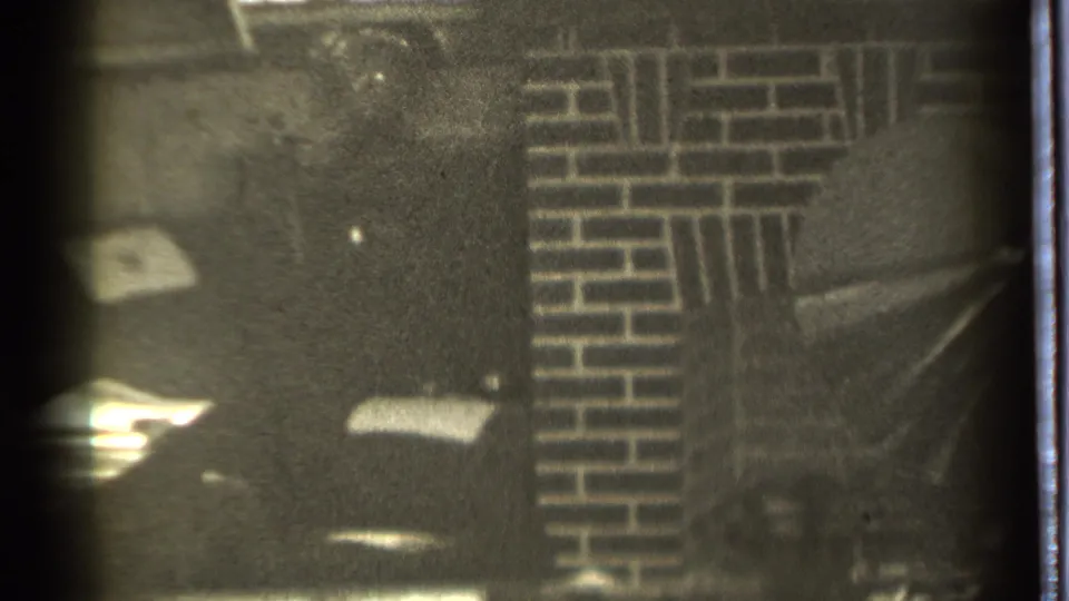 1939 Danielson home-movie frame showing a dim room, brick fireplace or wall, and a seated man partly visible at the bottom edge.