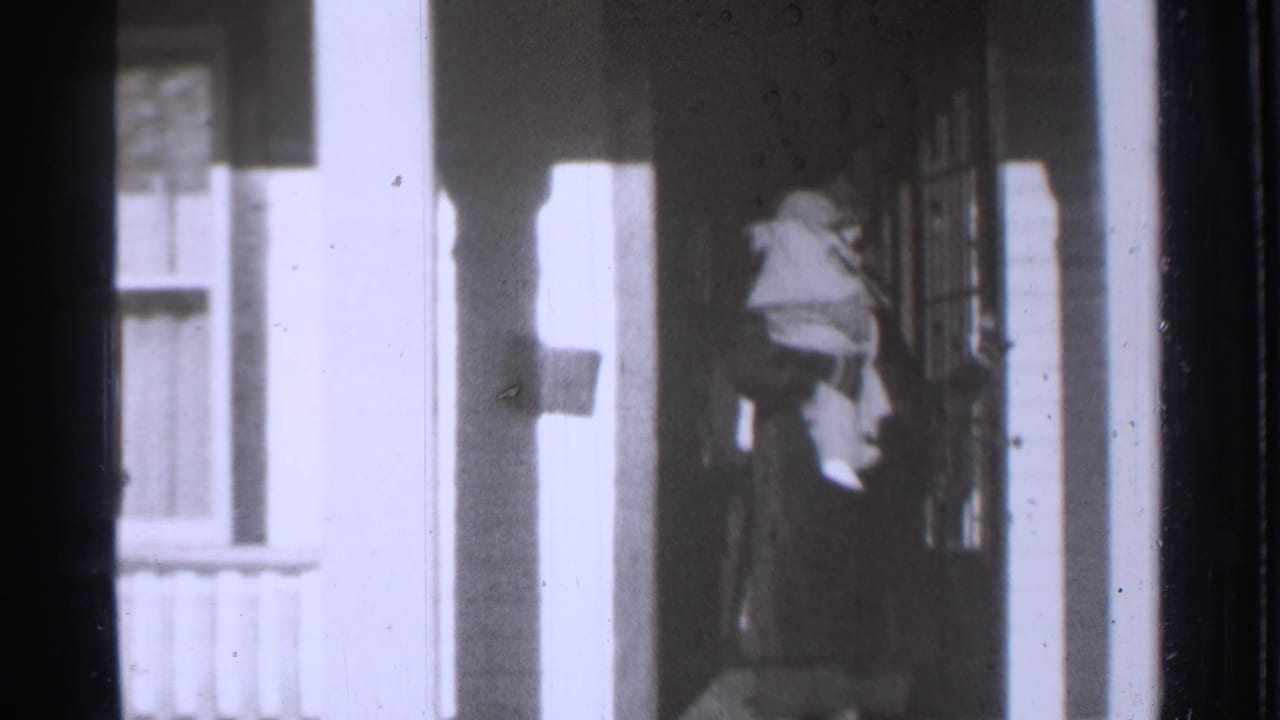 A figure in a light hat or hood stands near a porch doorway beside a window.