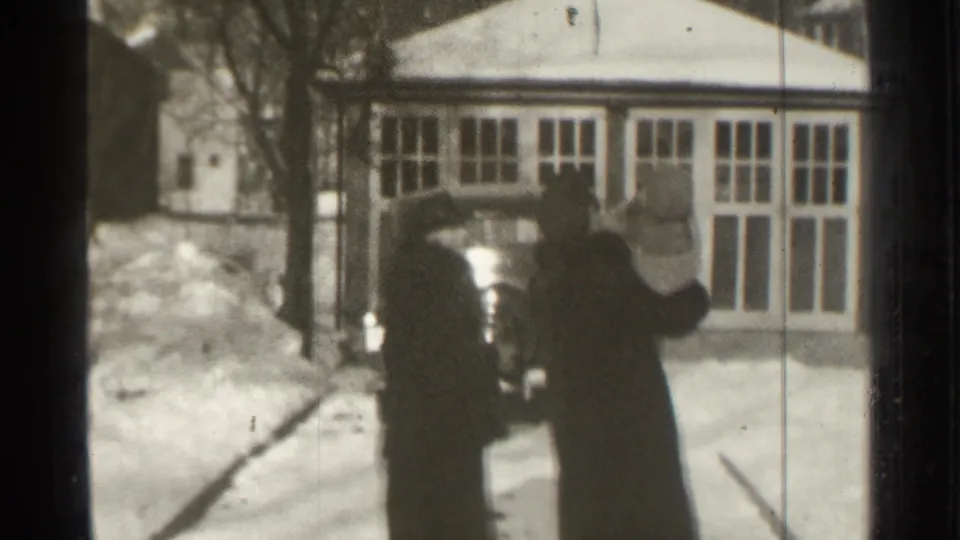 In Danielson, Connecticut in 1939, two adults in dark winter coats approach a parked car outside a windowed building.