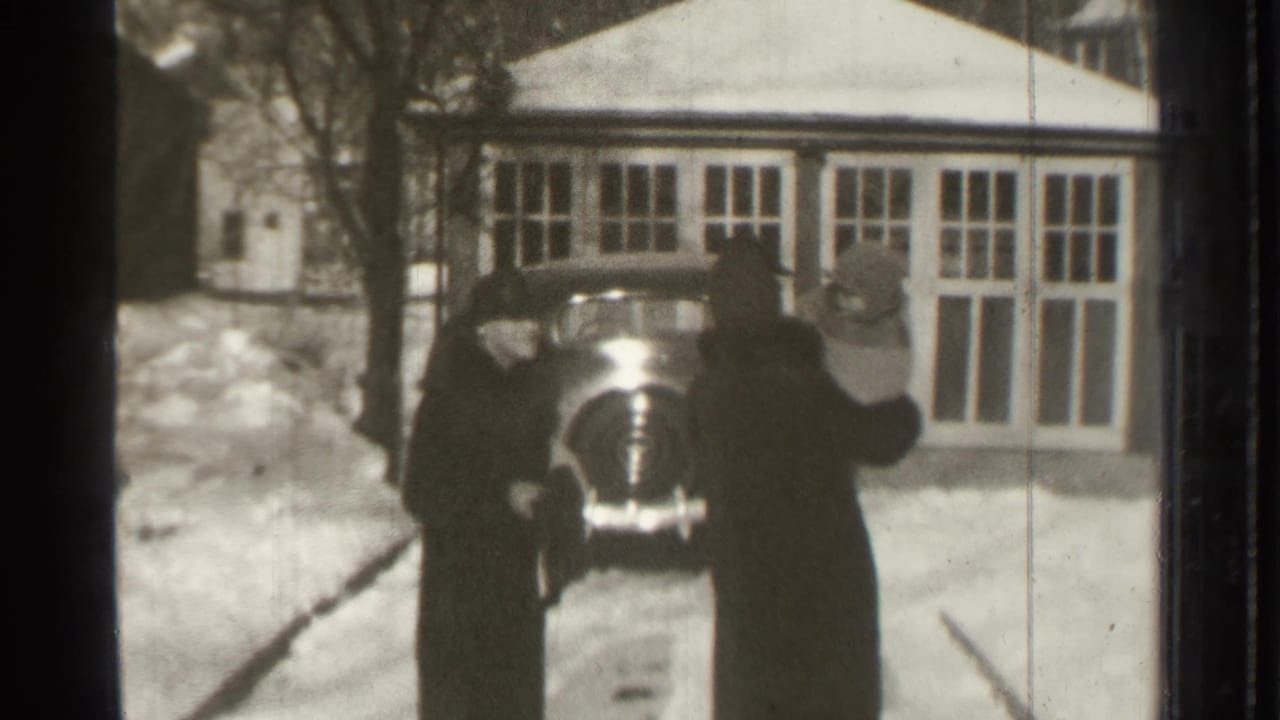 Two adults stand near the rear of a parked car on a snowy driveway.
