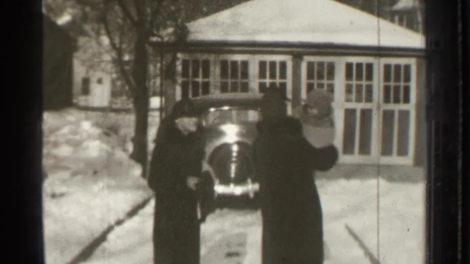Two coat-wearing adults stand by a parked car in a snowy Danielson driveway, with a multi-pane building facade behind them.