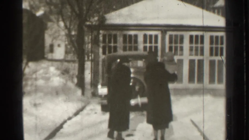 Two adults in heavy winter clothing stand near the rear of a dark car outside a snow-covered Danielson building facade.