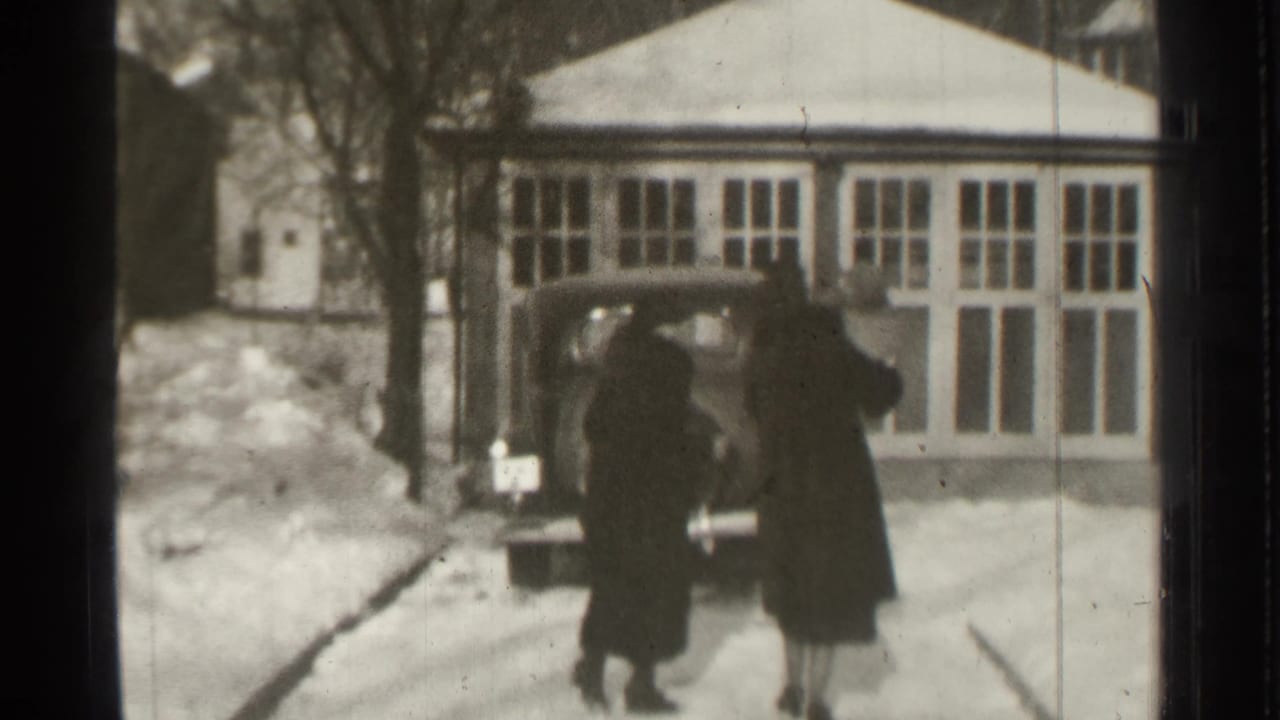 Two adults in dark coats walk toward a parked vintage car beside snowbanks.
