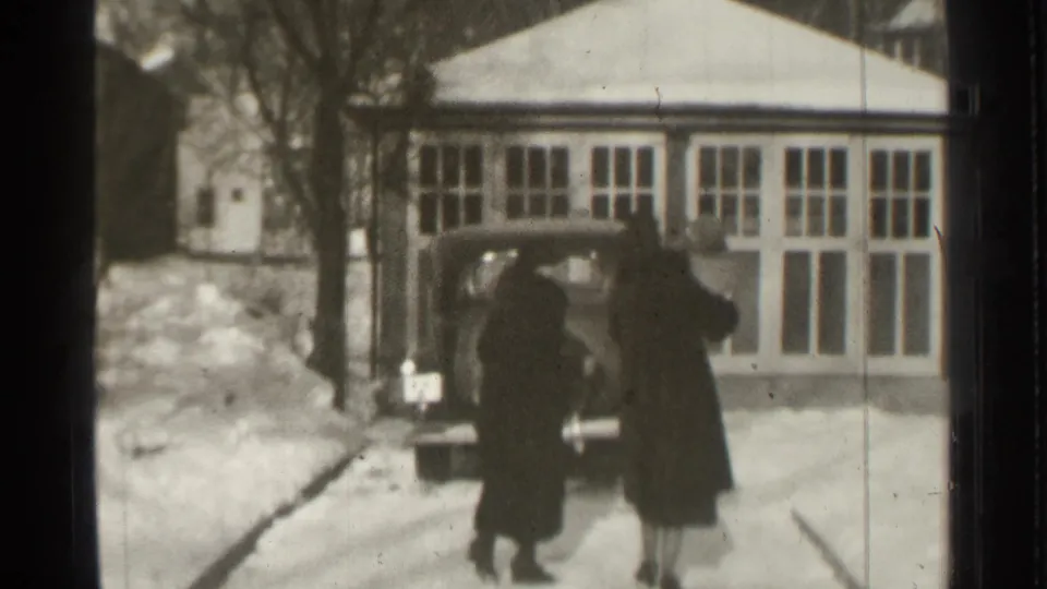 In 1939 Danielson, Connecticut, two adults in long dark coats approach a rounded vintage car on a snowy residential driveway.