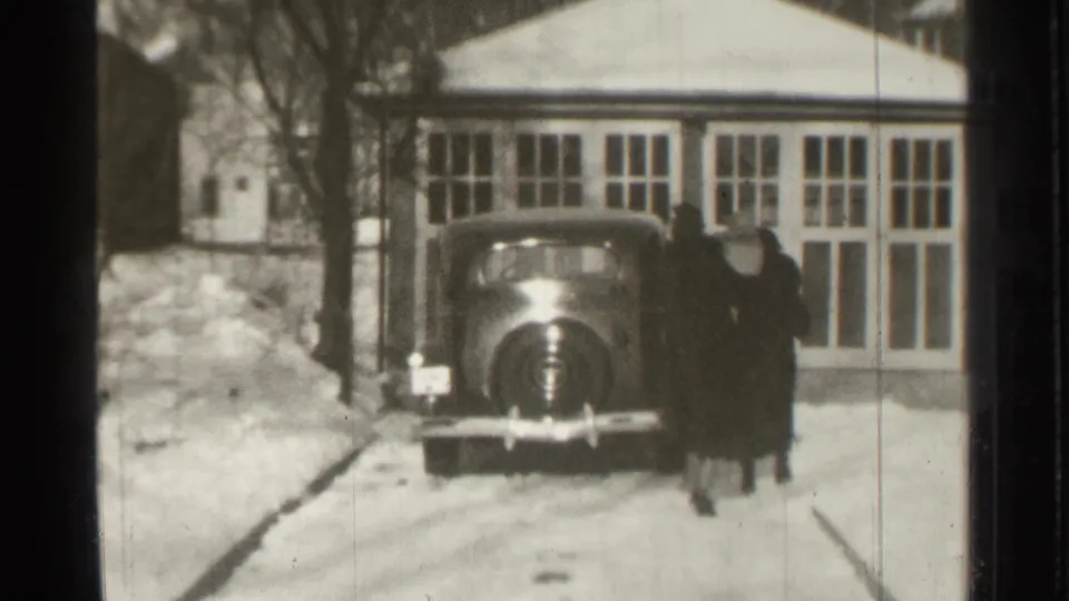 A dark 1930s-style car sits in the snowy driveway while two adults in long coats stand near its side in the home-movie frame.