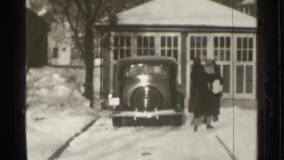 The pair in dark coats move along the side of the parked vintage car, with snowbanks and large windows behind them in Danielson, Connecticut.