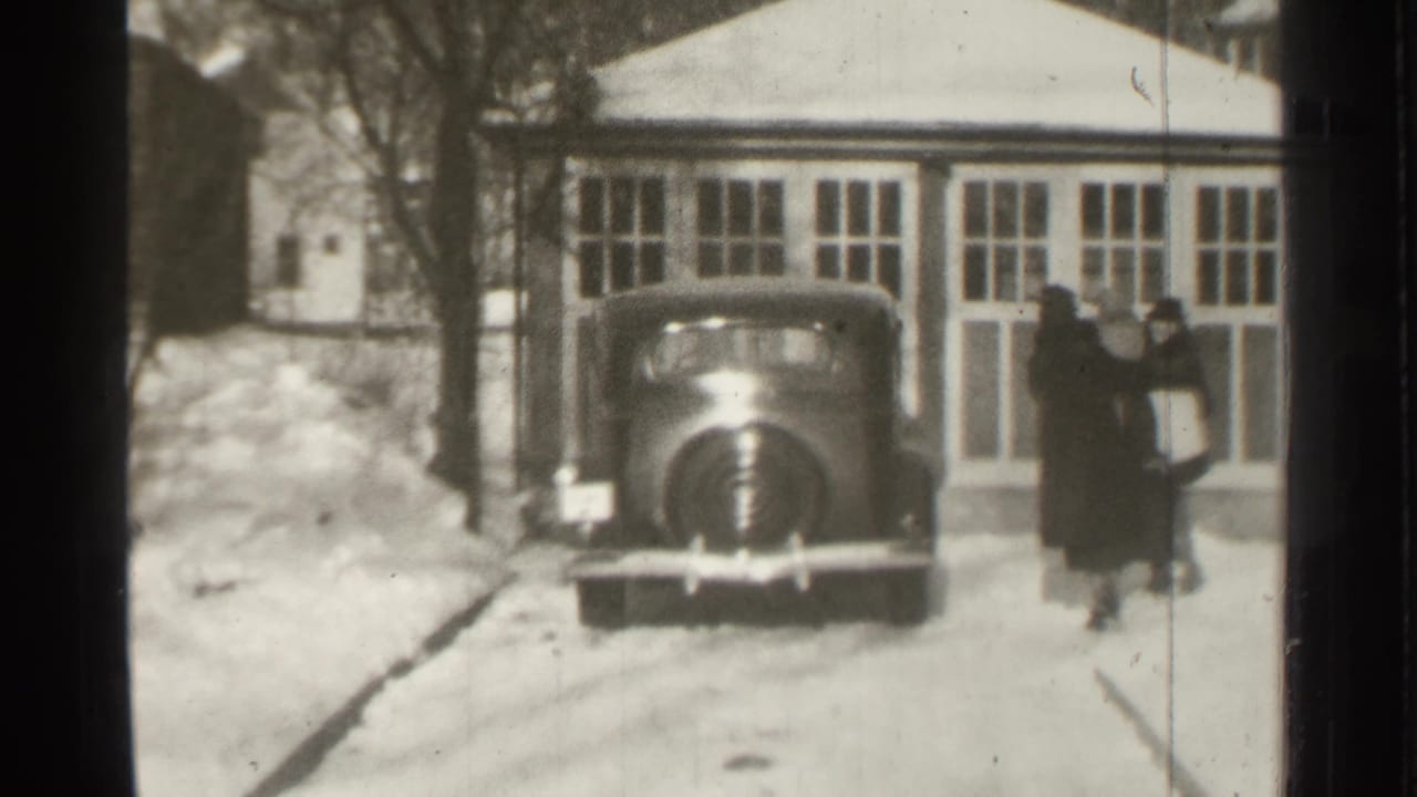Two adults walk beside a parked 1930s car in a snowy driveway near a garage.