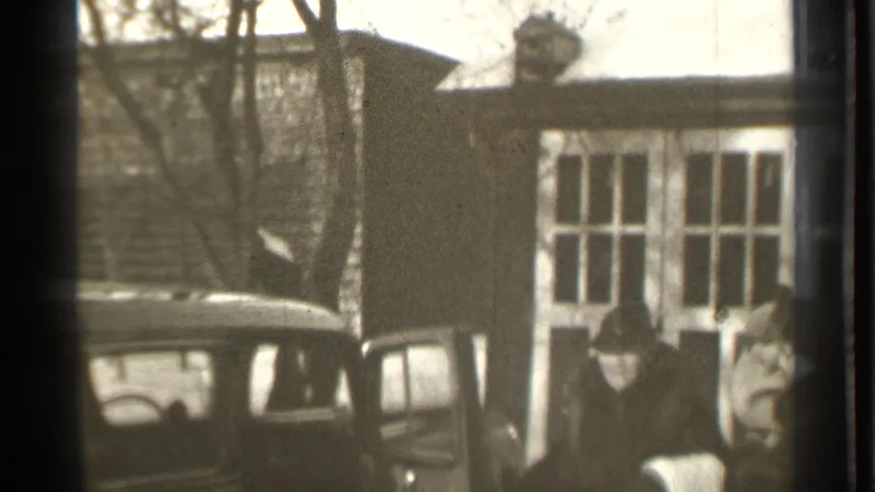 Adults in winter coats gather around the open door of a 1930s automobile outside a snowy Danielson residence.