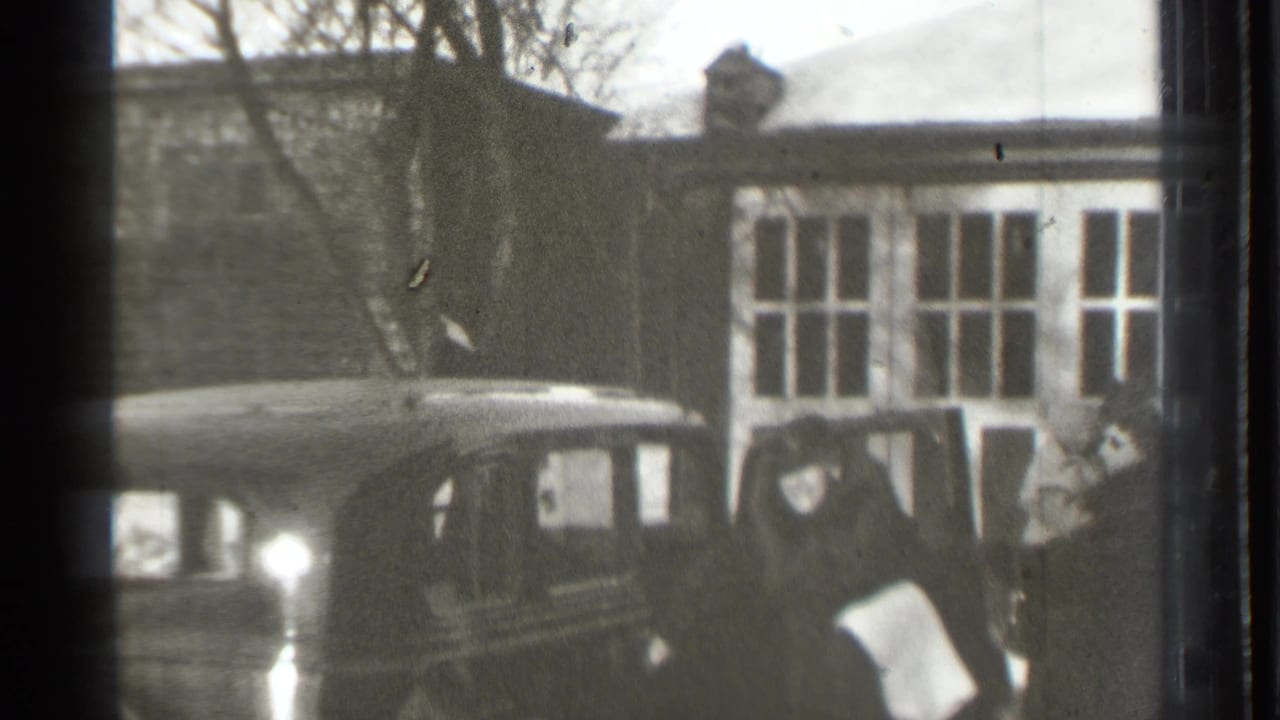 Dark sedan in front of a house as a woman holding an infant stands nearby.