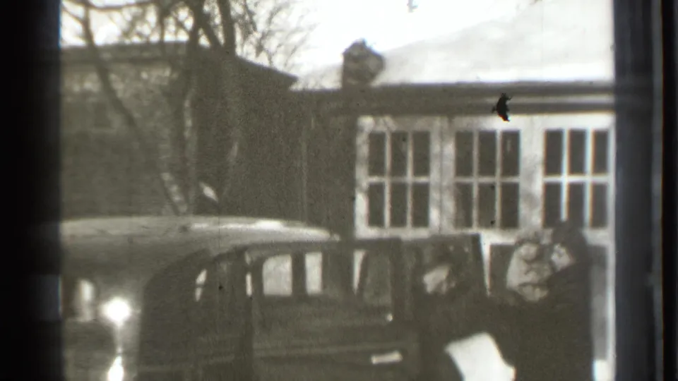 1939 Danielson home-movie frame with a boxy sedan and a woman holding an infant near a residential facade.