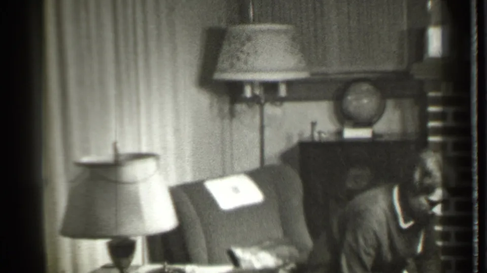 1939 Danielson, Connecticut home-movie frame showing a woman in a robe beside an armchair, with lamps, curtains, and a cabinet behind her.