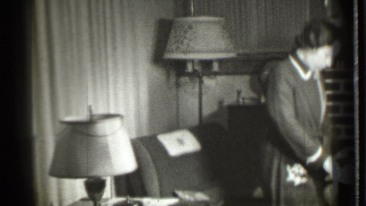 Woman standing beside a chair near a brick fireplace in a furnished room.