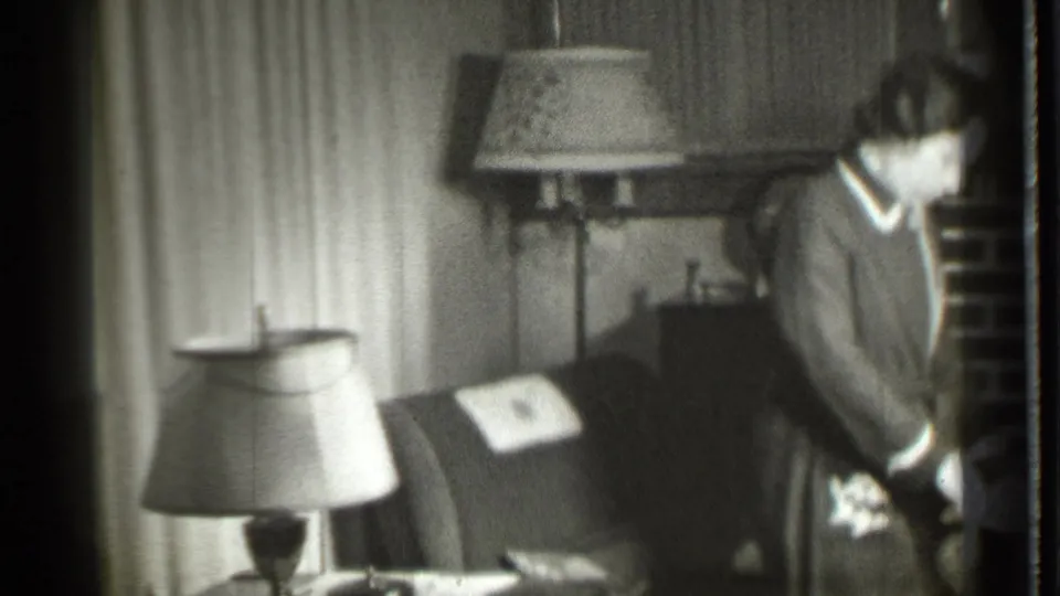 The woman stands near the right edge of a 1939 Danielson living room frame, beside an armchair, lamps, cabinet, globe, and brick fireplace.