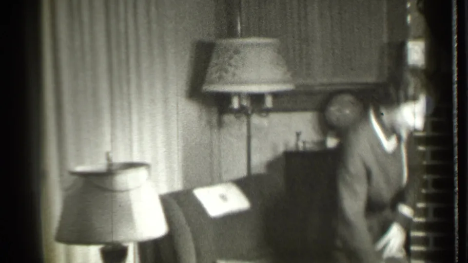 A woman in a grey and white robe stands beside a dark chair in a Danielson, Connecticut living room, with lamps, curtains, and fireplace details visible.