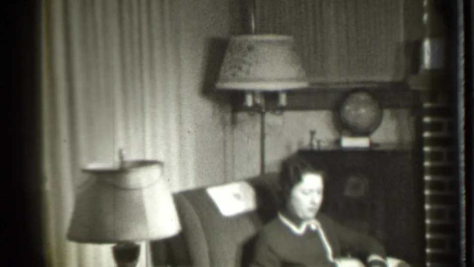 1939 home-movie frame from Danielson showing the woman seated in a dark armchair, framed by a floor lamp, cabinet, globe, and curtains.