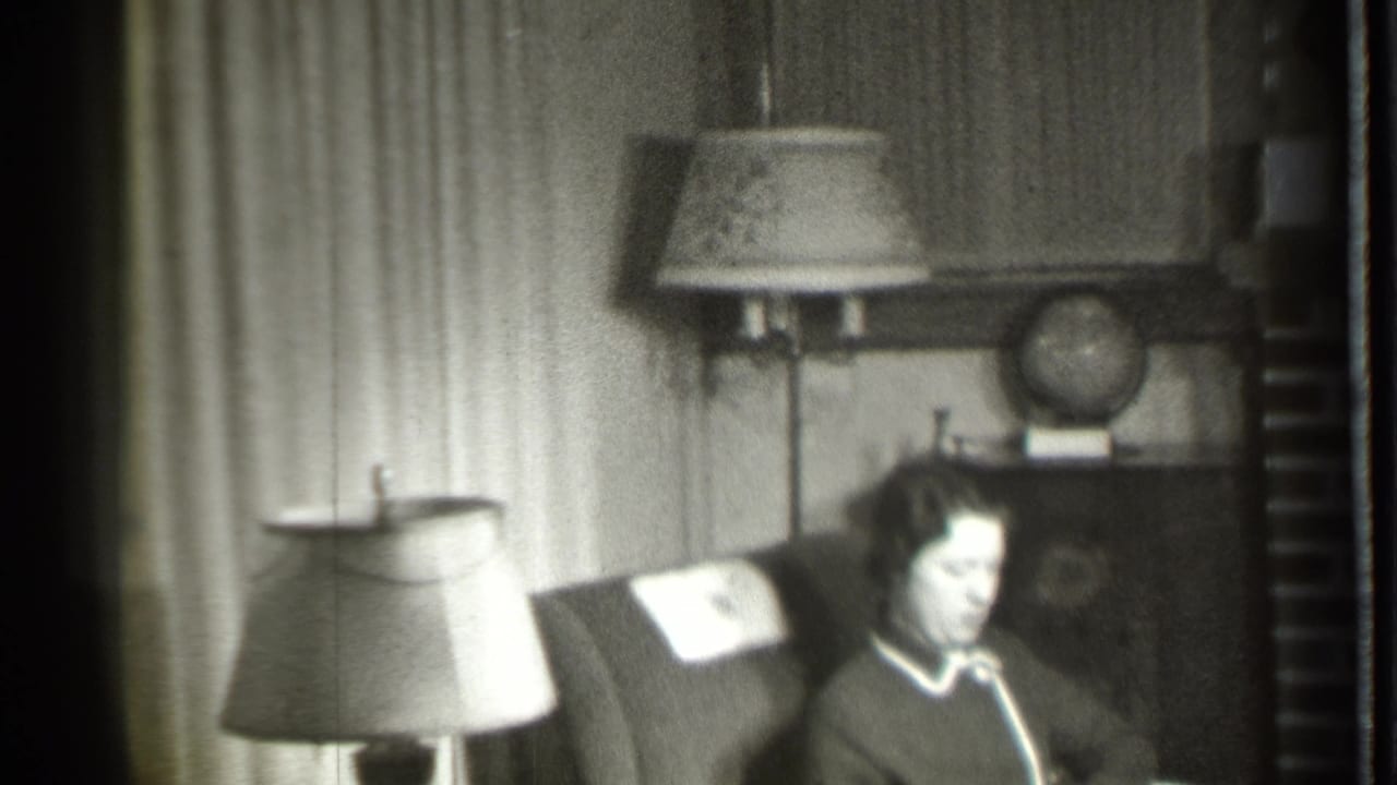 In a 1939 Danielson, Connecticut home interior, a woman sits in an upholstered armchair with lamps, curtains, a cabinet, and globe behind her.