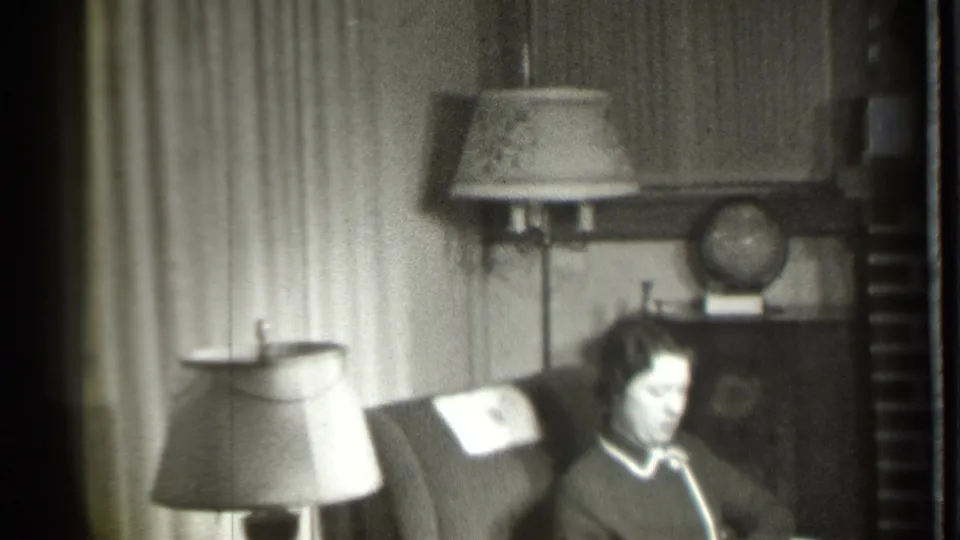In a 1939 Danielson, Connecticut home interior, a woman sits in an upholstered armchair with lamps, curtains, a cabinet, and globe behind her.