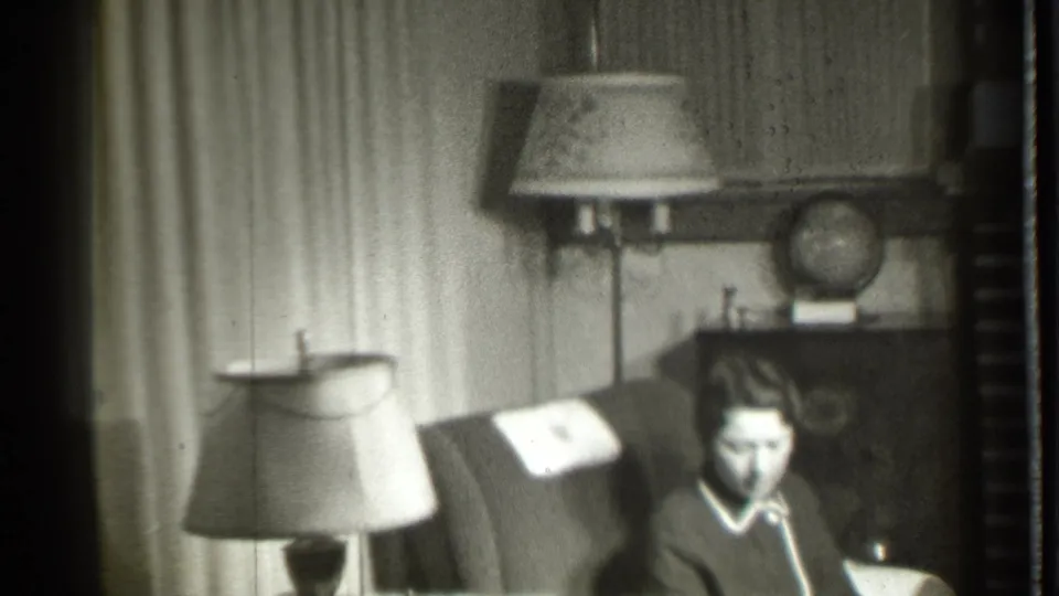 The woman sits low in the armchair in the Danielson living room, with a shaded lamp, curtains, cabinet, and globe visible in soft 8mm focus.