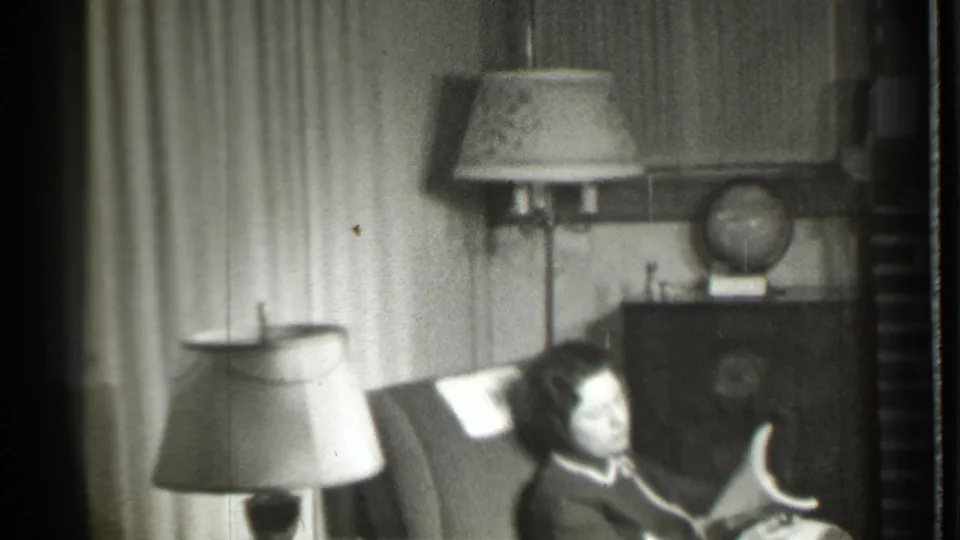The seated woman appears to handle a magazine or paper while sitting in a 1939 Danielson living room with lamps, curtains, cabinet, and globe.