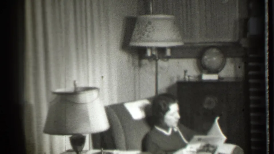 A woman reads a magazine in the 1939 Danielson home interior, seated beside a table lamp with a floor lamp, curtains, cabinet, and globe nearby.