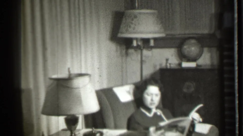 In a 1939 Danielson, Connecticut home interior, a woman sits by a lamp reading a magazine with curtains and a globe behind her.