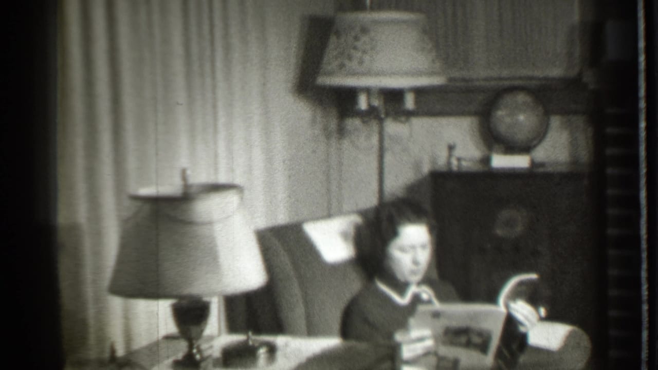 Woman holds an open magazine while seated near lamps and curtains.