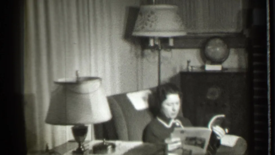 The woman remains seated in the Danielson living room, holding an open magazine with a table lamp in the foreground and a globe behind her.