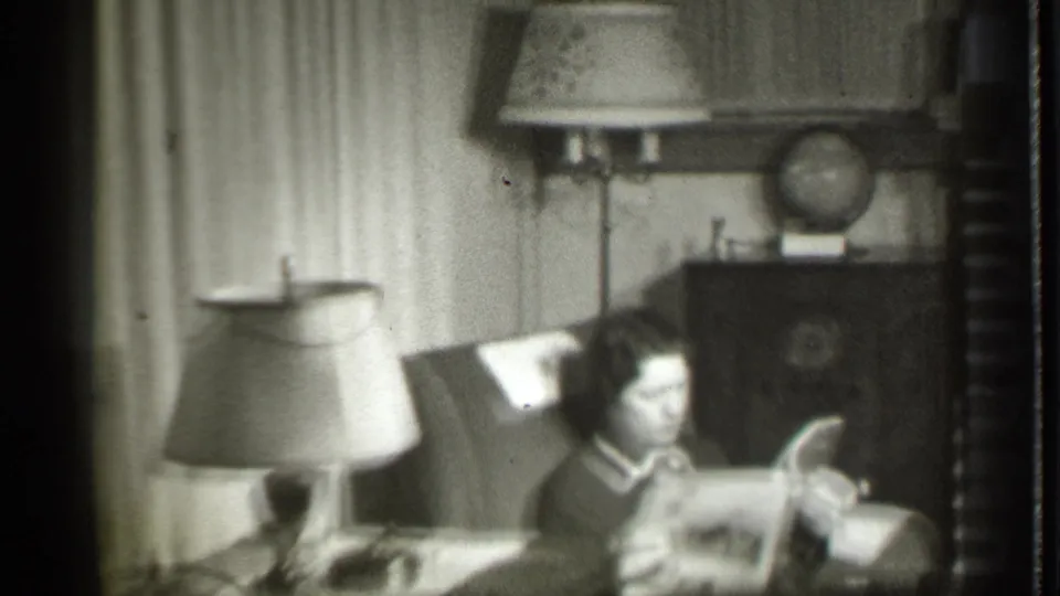 A woman in the 1939 home-movie frame looks down at a magazine or papers while seated among lamps, curtains, and a globe.