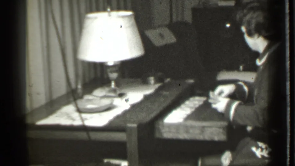 The woman appears at a desk or table in the private Danielson interior, handling papers beneath a bright lamp near curtains and dark furniture.