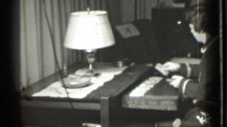 The woman reaches across a wooden table toward playing cards in a dim Danielson interior from 1939 home-movie footage.