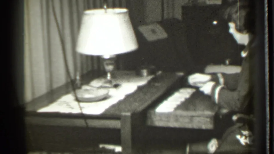 A woman remains seated beside a lamp and card layout, seen in black-and-white 1939 Danielson home-movie footage.