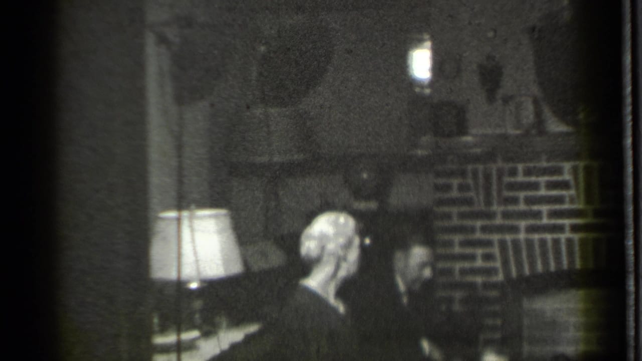 Two adults seated near a brick fireplace and table lamp in a dark living room.