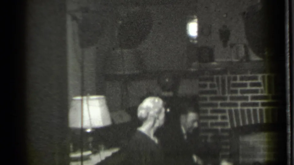 Danielson, Connecticut 1939 home-movie frame showing two adults seated in shadow near a brick fireplace and lit table lamp.