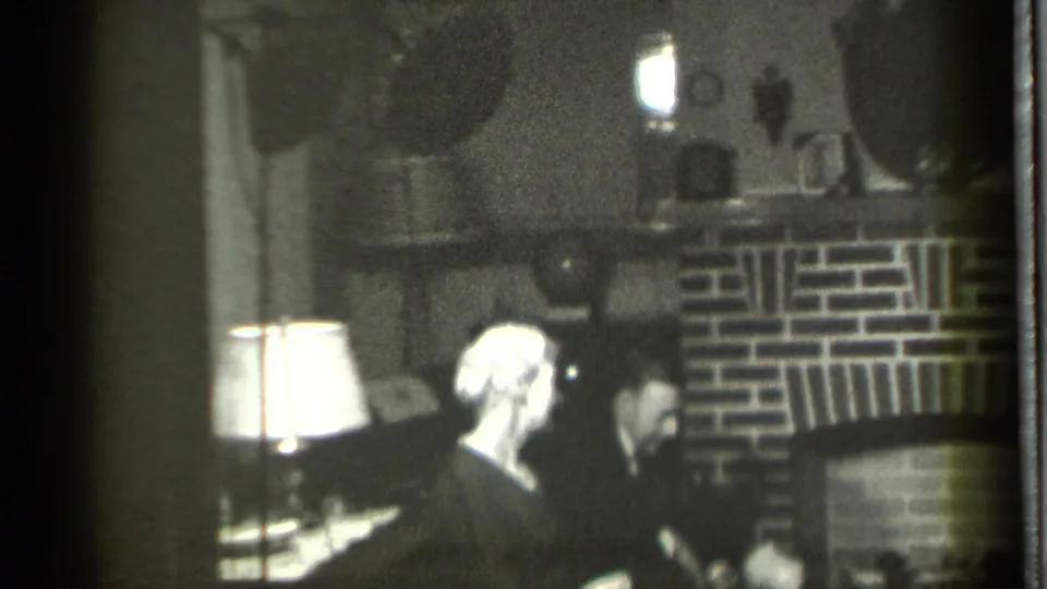 A 1939 Danielson home interior shows adults seated near a brick fireplace, with a white-shaded lamp providing the main light.
